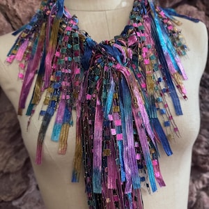 May include: A colorful, handmade necklace with a unique design. The necklace features a variety of ribbons in shades of pink, blue, purple, and gold. The ribbons are woven and knotted together, creating a textured and eye-catching accessory.