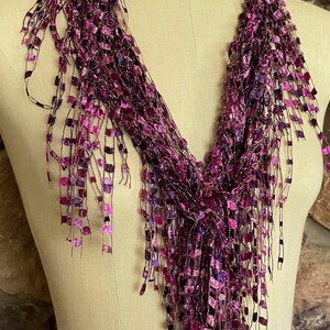 May include: A purple scarf with a fringed design. The scarf is draped over a mannequin and features a mix of dark and light purple squares and strands. The scarf has a shimmering, textured appearance.