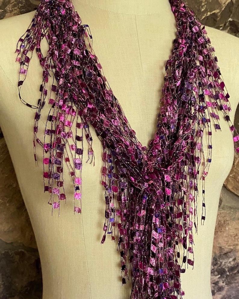 May include: A purple scarf with a fringed design. The scarf is draped over a mannequin and features a mix of dark and light purple squares and strands. The scarf has a shimmering, textured appearance.