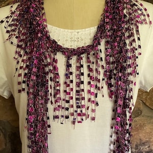 May include: A purple and pink scarf with a fringed design. The scarf is made of a mesh material with small, square accents in shades of purple and pink. The scarf is draped over a white shirt.