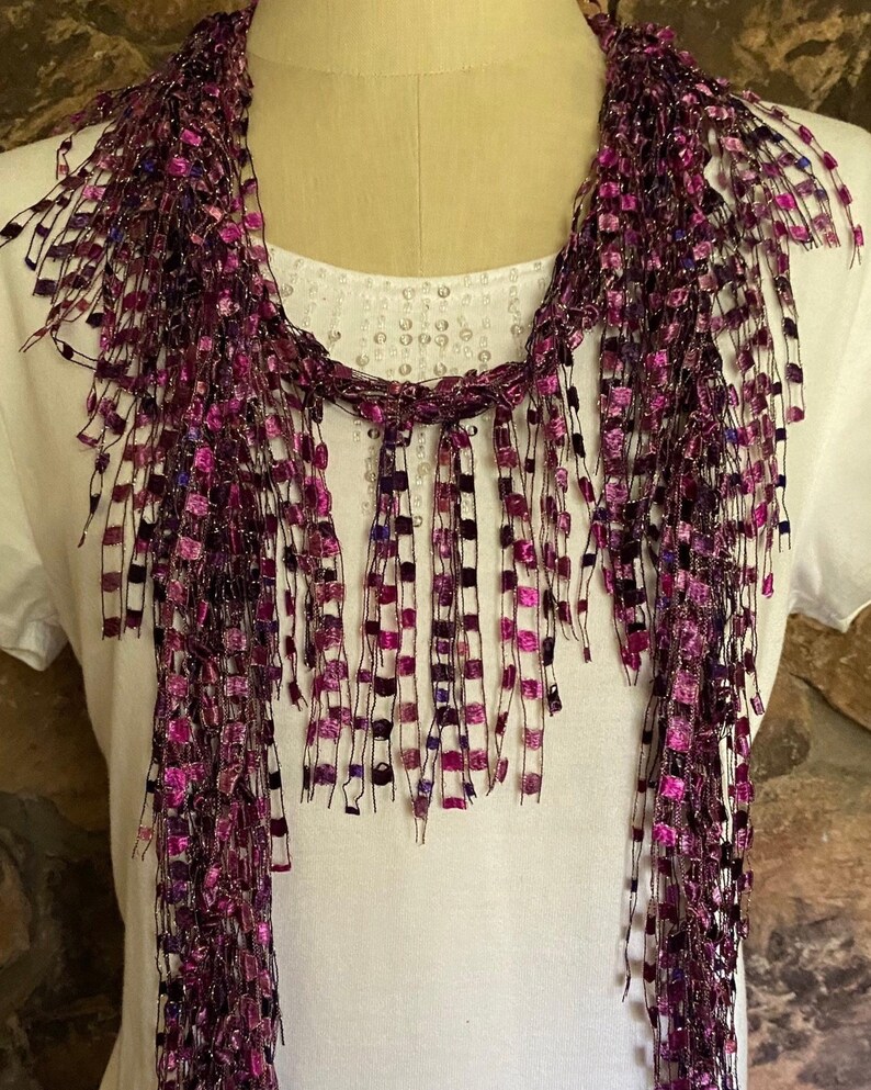 May include: A purple and pink scarf with a fringed design. The scarf is made of a mesh material with small, square accents in shades of purple and pink. The scarf is draped over a white shirt.