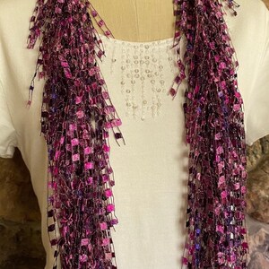 May include: A long, fringed scarf in shades of purple and pink. The scarf is made of a mesh-like material with small, square accents. The scarf is draped over a white shirt with a beaded design.