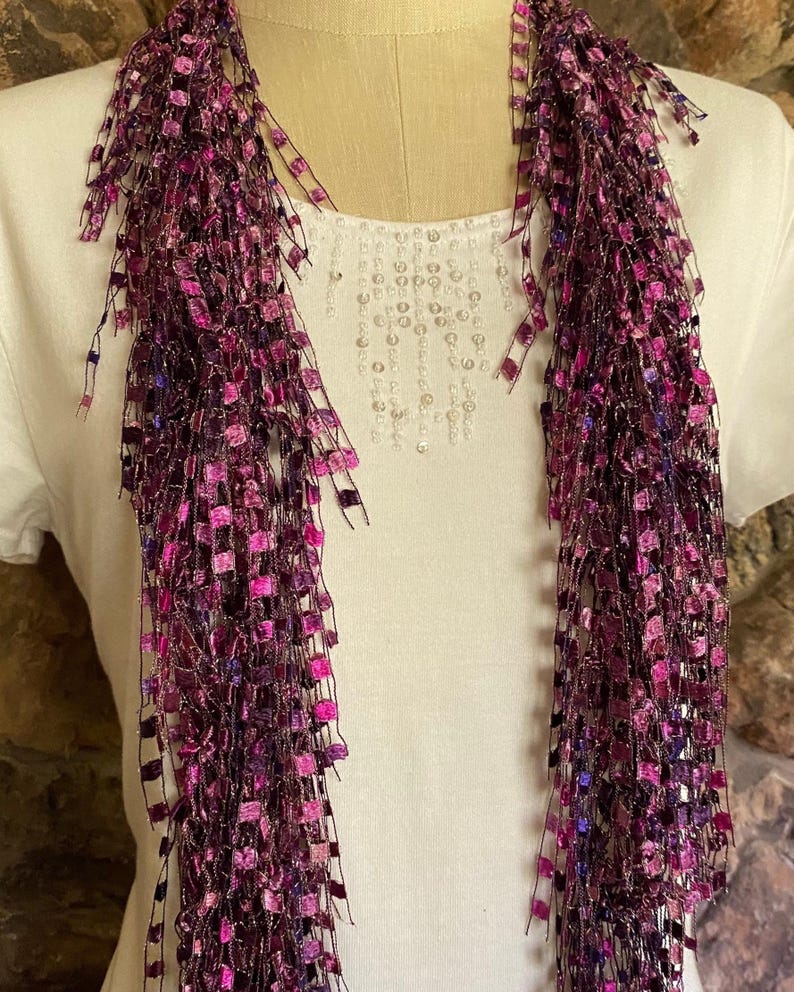 May include: A long, fringed scarf in shades of purple and pink. The scarf is made of a mesh-like material with small, square accents. The scarf is draped over a white shirt with a beaded design.