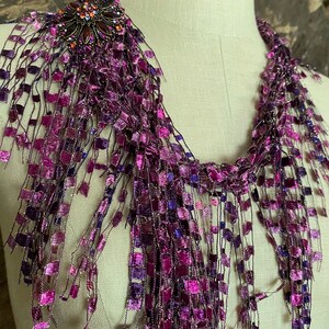 May include: A purple and magenta scarf with a decorative brooch. The scarf is made of a woven material with a fringe of square-shaped accents. The brooch is dark metal with orange and purple gemstones.