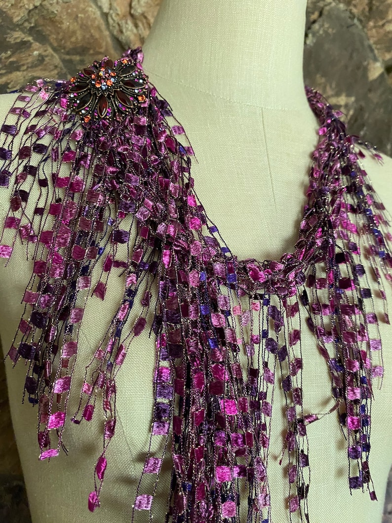 May include: A purple and magenta scarf with a decorative brooch. The scarf is made of a woven material with a fringe of square-shaped accents. The brooch is dark metal with orange and purple gemstones.