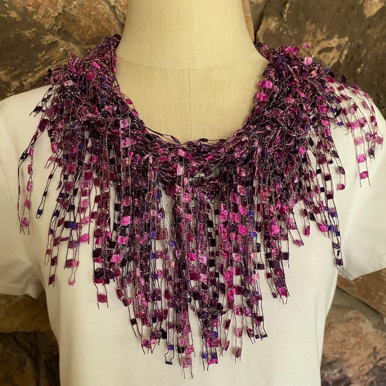 May include: A purple and pink crocheted fringe necklace with a square pattern. The necklace is on a white mannequin.