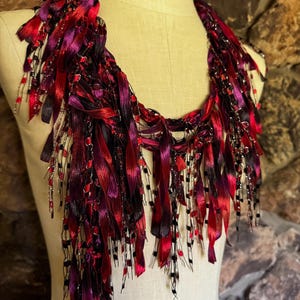 May include: A handmade necklace with a vibrant color palette of red, purple, and black. The necklace features long, flowing ribbons and beaded strands, creating a layered and textured effect. The design is displayed on a mannequin.