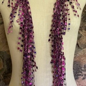 May include: A long, fringed scarf in shades of purple, magenta, and blue is draped on a mannequin. The scarf features a square, textured design with metallic threads, creating a shimmering effect. The scarf's fringe hangs down, adding movement.
