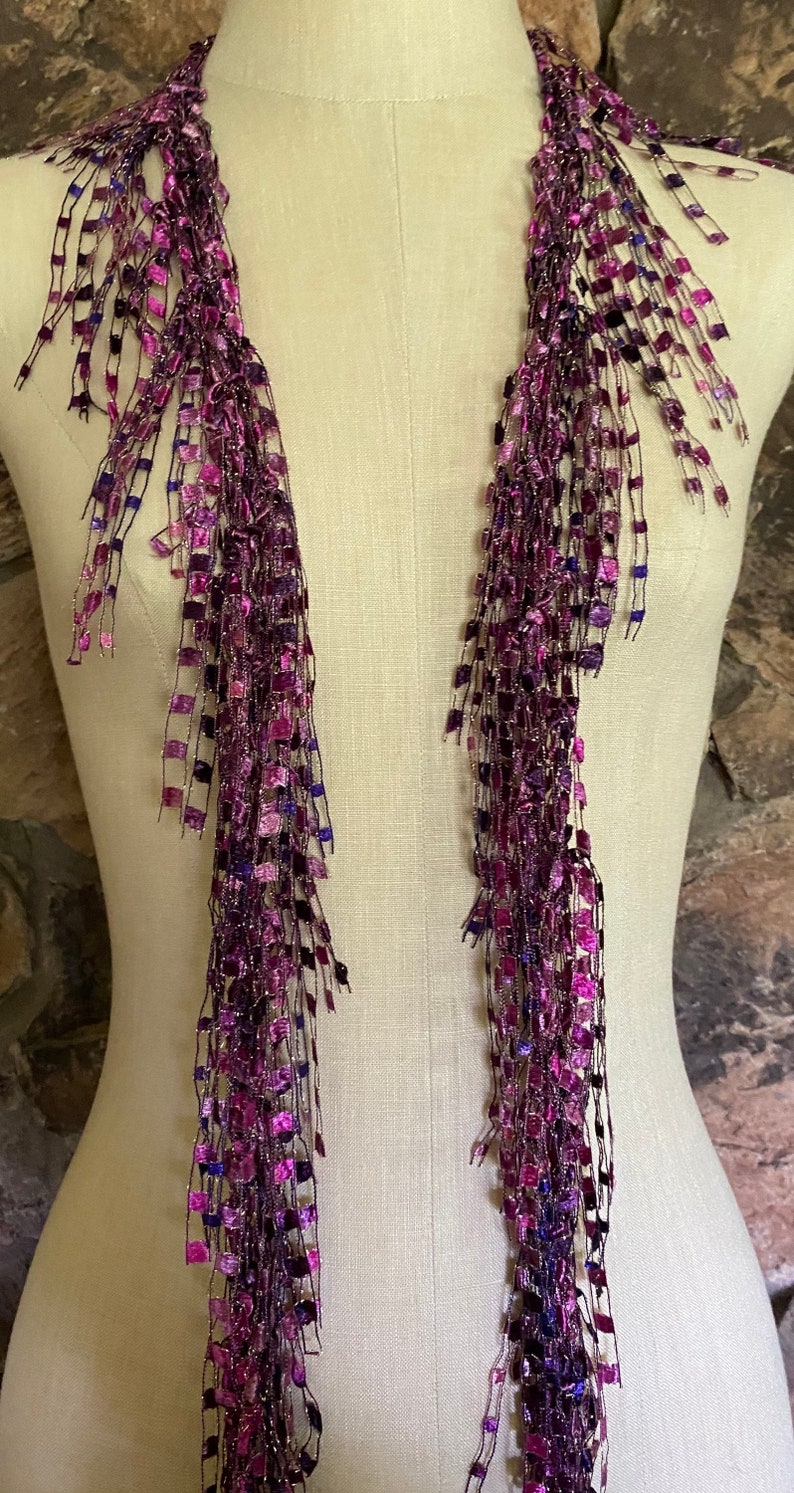 May include: A long, fringed scarf in shades of purple, magenta, and blue is draped on a mannequin. The scarf features a square, textured design with metallic threads, creating a shimmering effect. The scarf's fringe hangs down, adding movement.
