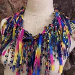 May include: A colorful, handmade necklace with a vibrant array of ribbons in shades of blue, pink, yellow, and green. The necklace features a layered design with beaded accents, creating a unique and eye-catching accessory.