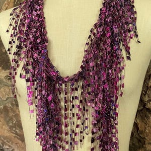 May include: A purple and pink scarf with a fringe design. The scarf is made of a soft, shimmering material and is draped over a mannequin. The fringe is made of small, square-shaped pieces.