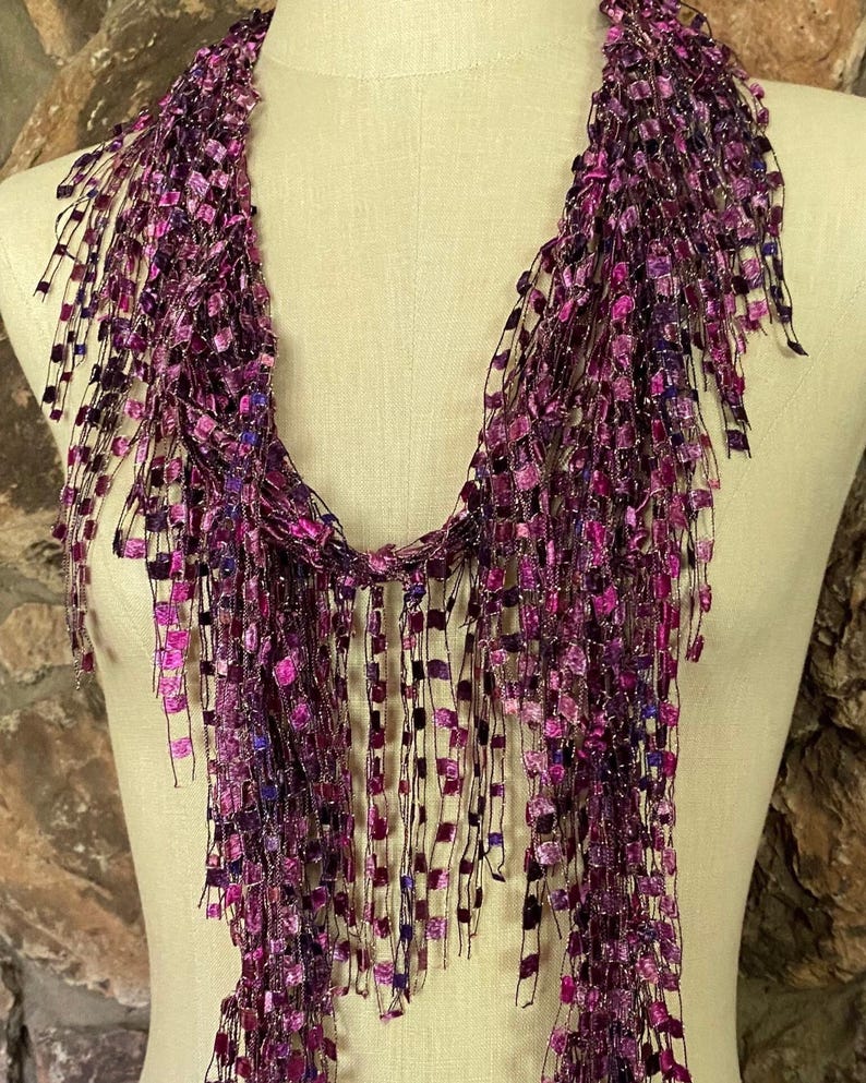 May include: A purple and pink scarf with a fringe design. The scarf is made of a soft, shimmering material and is draped over a mannequin. The fringe is made of small, square-shaped pieces.