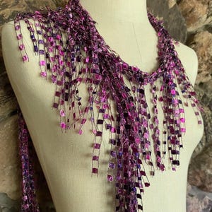May include: A purple scarf with a fringe design. The scarf is draped over a mannequin. The fringe is made of small, square-shaped pieces in shades of purple and pink. The background is a stone wall.