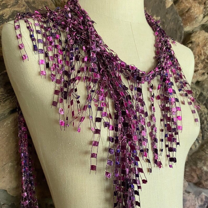 May include: A purple scarf with a fringe design. The scarf is draped over a mannequin. The fringe is made of small, square-shaped pieces in shades of purple and pink. The background is a stone wall.