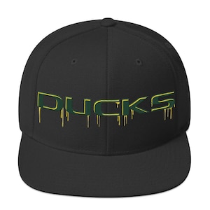 Op de afbeelding: Zwarte baseballcap met het woord "DUCKS" in groen en geel. De letters hebben een druipend effect, met gele druppels. De cap heeft een platte klep en een gestructureerde pasvorm.