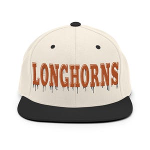 Könnte beinhalten: Cremefarbene Baseballkappe mit flachem Schirm und dem Wort "LONGHORNS" in orangefarbenen, tropfenden Buchstaben. Die Kappe hat einen schwarzen Schirm und einen schwarzen Knopf oben. Sportliches Accessoire.