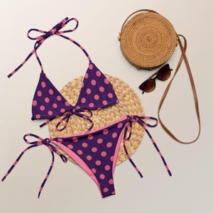 May include: A purple and pink polka dot bikini with adjustable straps. The bikini is on a woven straw mat with a straw bag and sunglasses.
