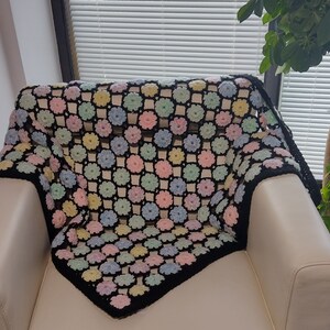 May include: A crocheted throw blanket with a black background and colourful flower designs. The blanket is draped over a white armchair.