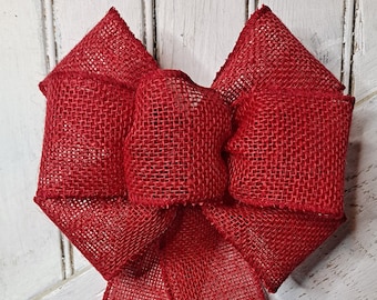May include: A large red burlap bow with two long tails. The bow is tied in a classic shape with a center loop. The tails are long and flowing, and the burlap is a coarse, textured fabric.