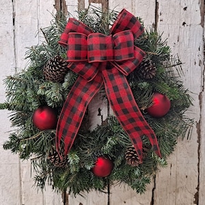 May include: A Christmas wreath with evergreen branches, pinecones, and red ornaments. A large red and black buffalo plaid bow is centered at the top. The wreath is displayed against a white, weathered wooden background.