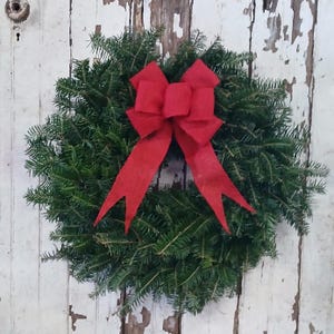 May include: A circular wreath made of dense, dark green evergreen needles. A large, bright red burlap bow with two long, pointed tails is centered at the top of the wreath, providing a bold contrast against the natural green foliage.