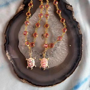 Könnte beinhalten: Zwei goldene Halsketten mit rosa Edelstein-Gliedern und Schildkröten-Anhängern. Die Schildkröten-Anhänger sind weiß und rosa mit Blumen- und Muscheldesign. Die Halsketten werden auf einem grau-weißen Achatstein präsentiert.