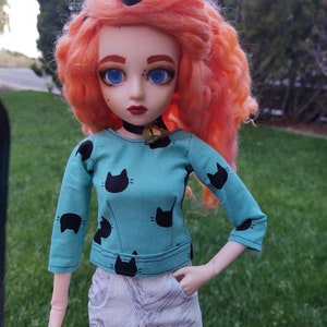 Pode incluir: Uma boneca com olhos azuis brilhantes, cabelo rosa e um laço preto. Ela está vestindo uma camisa turquesa com silhuetas de gatos pretos e calças brancas.