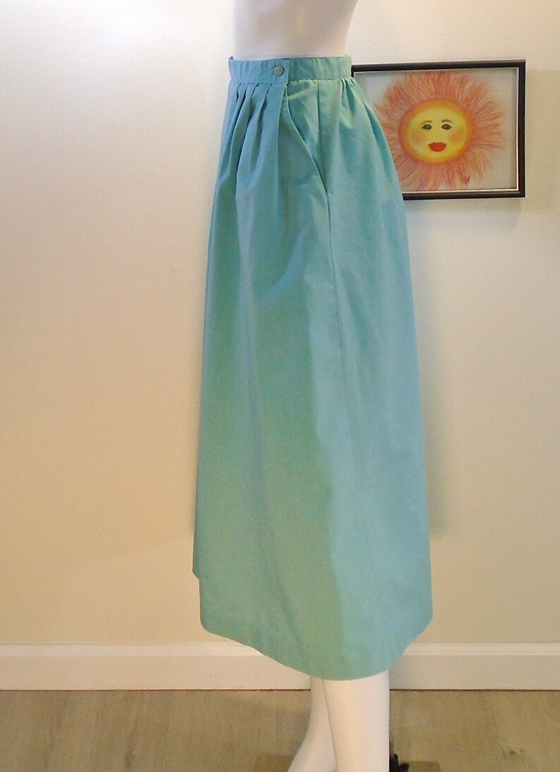 May include: A light blue midi skirt with a single button closure at the waist. The skirt has pleats at the front and a single pocket on the right side.