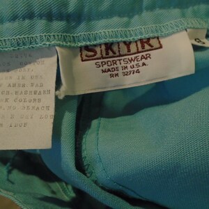 May include: A white tag with black text that reads "ROTY WM 52774 CONTON IN USA OF AMER. FAD CHACH WASHWARN DARK COLORS PANO BERACH 48-B DRY LOW" attached to a light blue garment with a brown tag that reads "SKYK SPORTSWEAR MADE IN U.S.A. RM 32774".