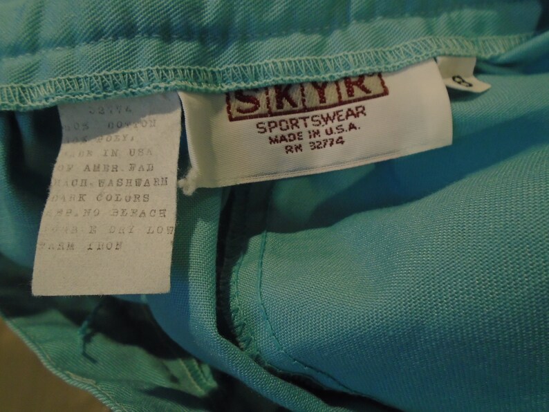 May include: A white tag with black text that reads "ROTY WM 52774 CONTON IN USA OF AMER. FAD CHACH WASHWARN DARK COLORS PANO BERACH 48-B DRY LOW" attached to a light blue garment with a brown tag that reads "SKYK SPORTSWEAR MADE IN U.S.A. RM 32774".