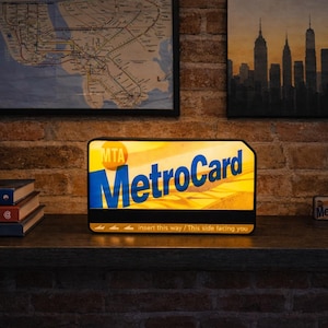 May include: A rectangular, illuminated sign designed to resemble a New York City MTA MetroCard. The sign features the blue and yellow MetroCard logo, the text 'MTA', and the instructional text 'insert this way / This side facing you' at the bottom.