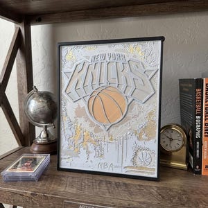 May include: A framed 3D wall art piece featuring the New York Knicks logo in white and tan tones. The design includes a central basketball graphic, textured white layers with gold accents, and the text New York Knicks and NBA printed on the front.