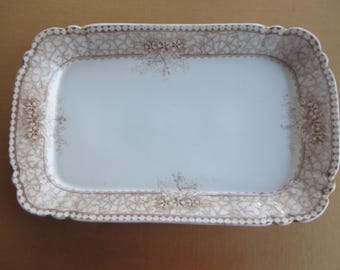 May include: Rectangular antique serving platter with a white center and a decorative border. The border features a brown floral and geometric pattern. The edges are scalloped. A vintage piece for serving food or display.