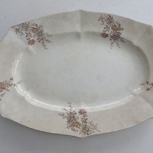 May include: An oval, off-white serving platter with a scalloped edge. The platter features floral designs in shades of brown and tan, adding a vintage aesthetic. The dish is ideal for serving food at a dinner party.