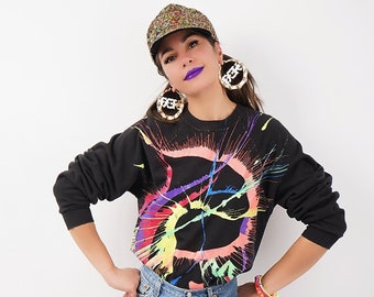 May include: A woman wearing a black sweatshirt with a colorful paint splatter design, a denim skirt, and multi-colored high-top sneakers. She is also wearing a sparkly green and gold baseball cap and large hoop earrings.