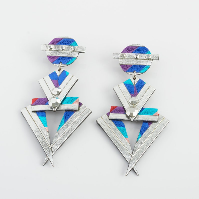 Może przedstawiać: Para srebrnych kolczyk&oacute;w z geometrycznymi kształtami i kolorowymi wzorami. Kolczyki mają centralny tr&oacute;jkąt z mniejszymi tr&oacute;jkątami i k&oacute;łkami wok&oacute;ł niego. Kolczyki są wykonane ze sk&oacute;ry i mają srebrne wykończenie.