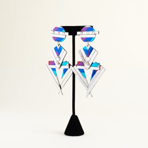 Może przedstawiać: Para srebrnych, geometrycznych kolczyk&oacute;w z biało-niebiesko-fioletowym wzorem w paski. Kolczyki mają duży, tr&oacute;jkątny kształt z mniejszym k&oacute;łkiem na g&oacute;rze.