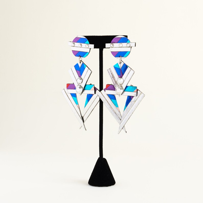 Może przedstawiać: Para srebrnych, geometrycznych kolczyk&oacute;w z biało-niebiesko-fioletowym wzorem w paski. Kolczyki mają duży, tr&oacute;jkątny kształt z mniejszym k&oacute;łkiem na g&oacute;rze.