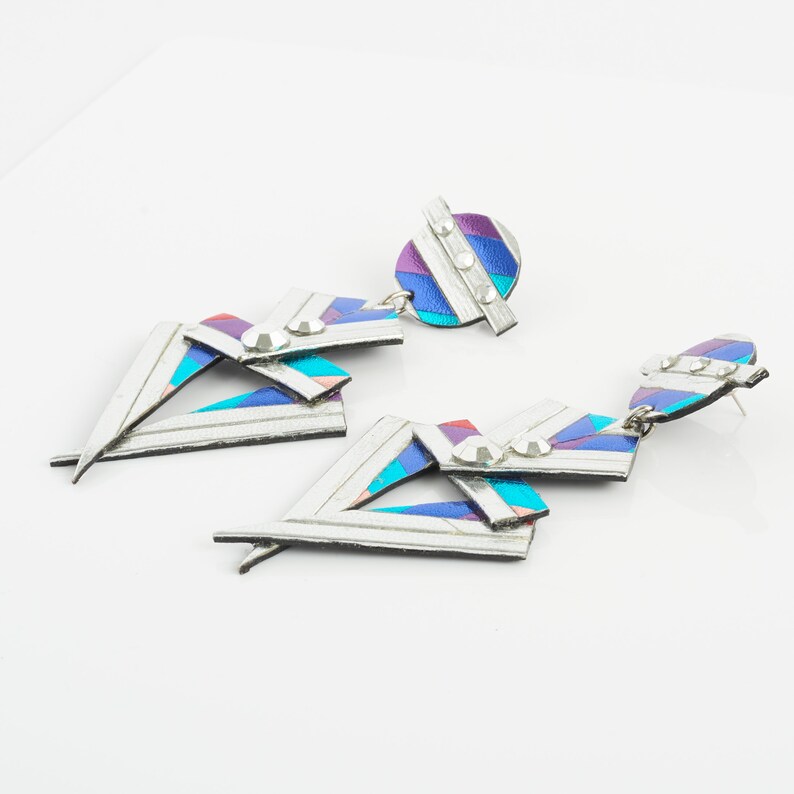 Może przedstawiać: Para srebrnych, geometrycznych kolczyk&oacute;w z wielokolorowym, tęczowym wzorem i przezroczystymi cyrkoniami.