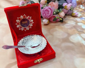 May include: A silver-colored, ornate bowl and spoon set inside a red velvet box. The box is open, revealing the detailed floral design on the bowl. The box has a gold clasp and a decorative label that says "Diwali".