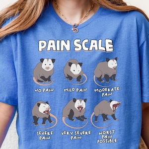 May include: A blue t-shirt featuring a pain scale with opossum illustrations. The text "PAIN SCALE" is at the top, with opossums depicting levels from "No Pain" to "Worst Pain Possible".