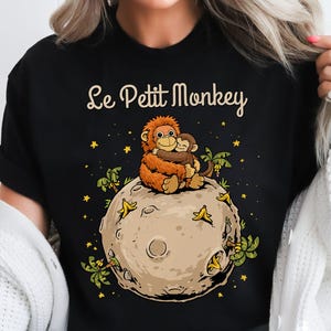 May include: Black t-shirt with the text "Le Petit Monkey" above a cartoon illustration of two monkeys hugging on a moon. The moon is tan with green and yellow accents, and the background is black with yellow stars.