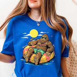 Może przedstawiać: Królewsko niebieska koszulka z kreskówkowym Bigfootem relaksującym się na kole do pływania z drinkiem. Bigfoot ma okulary przeciwsłoneczne, kwiatową girlandę i kąpielówki. Żółte słońce i niebieska woda w tle.