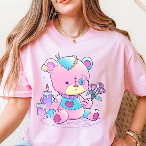 Whimsical light pink T-shirt featuring a colorful stitched voodoo teddy bear holding roses while sitting inside a pentagram surrounded by candles, ideal for fans of pastel goth, spooky-cute aesthetics, and alternative kawaii gifts.