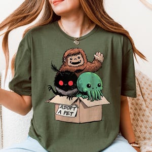 Whimsical olive green T-shirt featuring a cartoon Bigfoot, Mothman, and Cthulhu sitting inside a cardboard box labeled "ADOPT A PET," ideal for fans of cryptozoology, urban legends, and cute alternative-style graphic apparel.