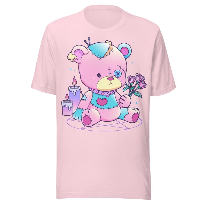 May include: Pink t-shirt with a cartoon image of a patched-up teddy bear holding a bouquet of pink roses. The bear is sitting on a pentagram with two candles behind it.