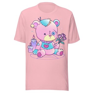 May include: A pink t-shirt with a cartoon image of a pink and blue stitched teddy bear holding a bouquet of pink roses. The bear is sitting on a pentagram with two purple candles.