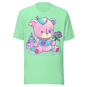 May include: A green t-shirt with a pink and blue patched teddy bear holding a bouquet of pink roses. The bear is sitting on a pentagram with two candles behind it.
