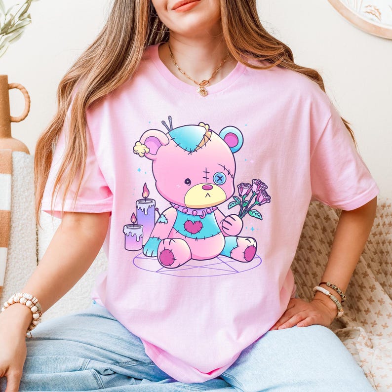 Whimsical light pink T-shirt featuring a colorful stitched voodoo teddy bear holding roses while sitting inside a pentagram surrounded by candles, ideal for fans of pastel goth, spooky-cute aesthetics, and alternative kawaii gifts.