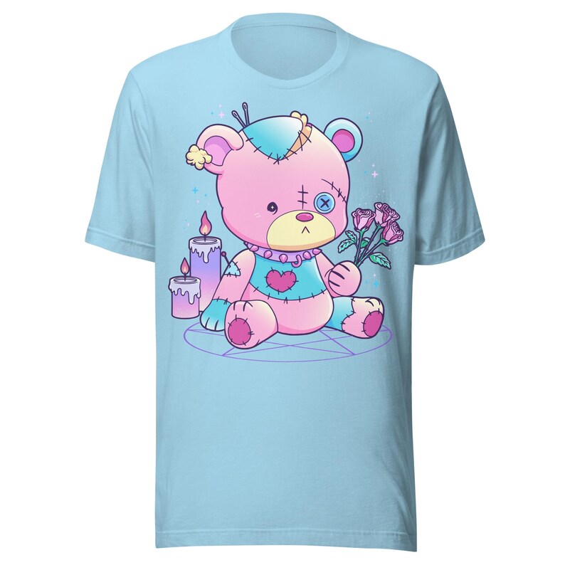 May include: A light blue t-shirt with a pink and blue stitched teddy bear holding a bouquet of pink roses. The bear is sitting on a pentagram with two purple candles behind it.